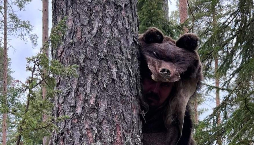 Karhuntaljaan pukeutunut mies kurkistaa puun takaa.
